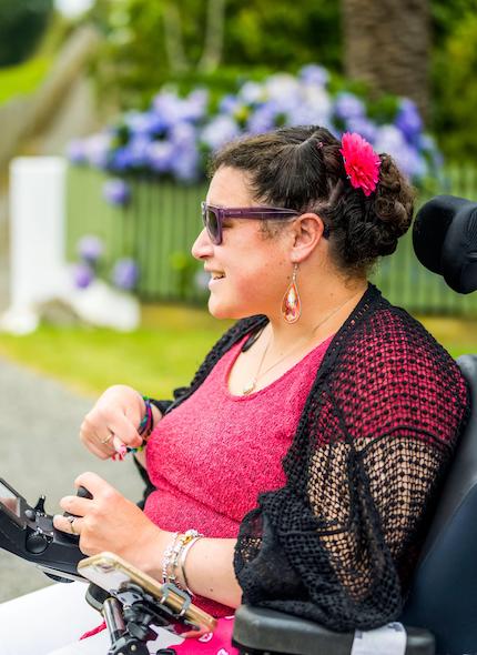 Smiling Māori woman who has Cerebral Palsy and is wearing fabulous earrings drives her motorised wheelchair past a park with hydrangeas.